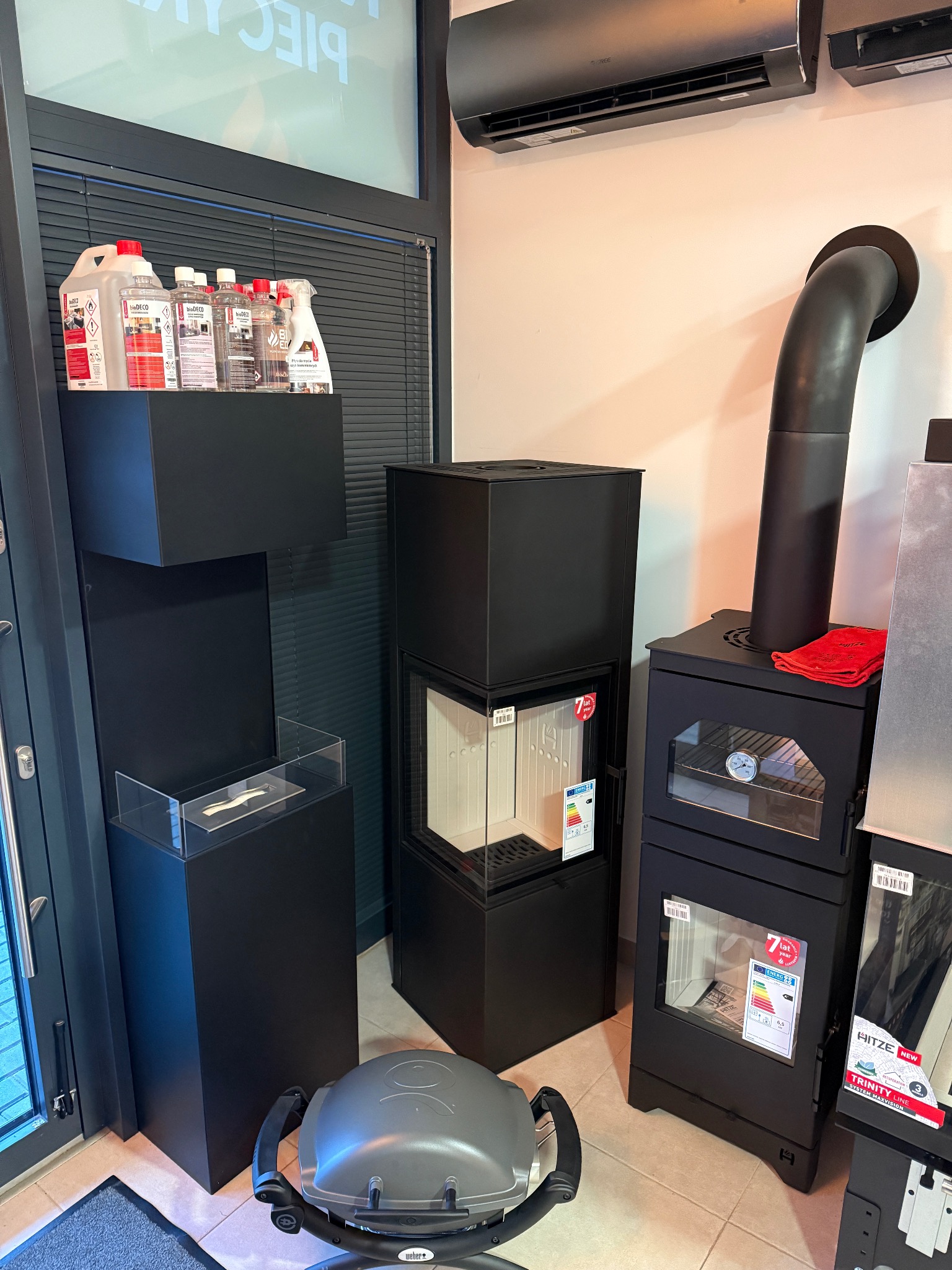 Ekspozycja w salonie z czarnymi kominkami i piecami, w tym piec z piekarnikiem i termometrem, akcesoria do czyszczenia i bio-kominek na postumencie, oraz grill Weber na podłodze.