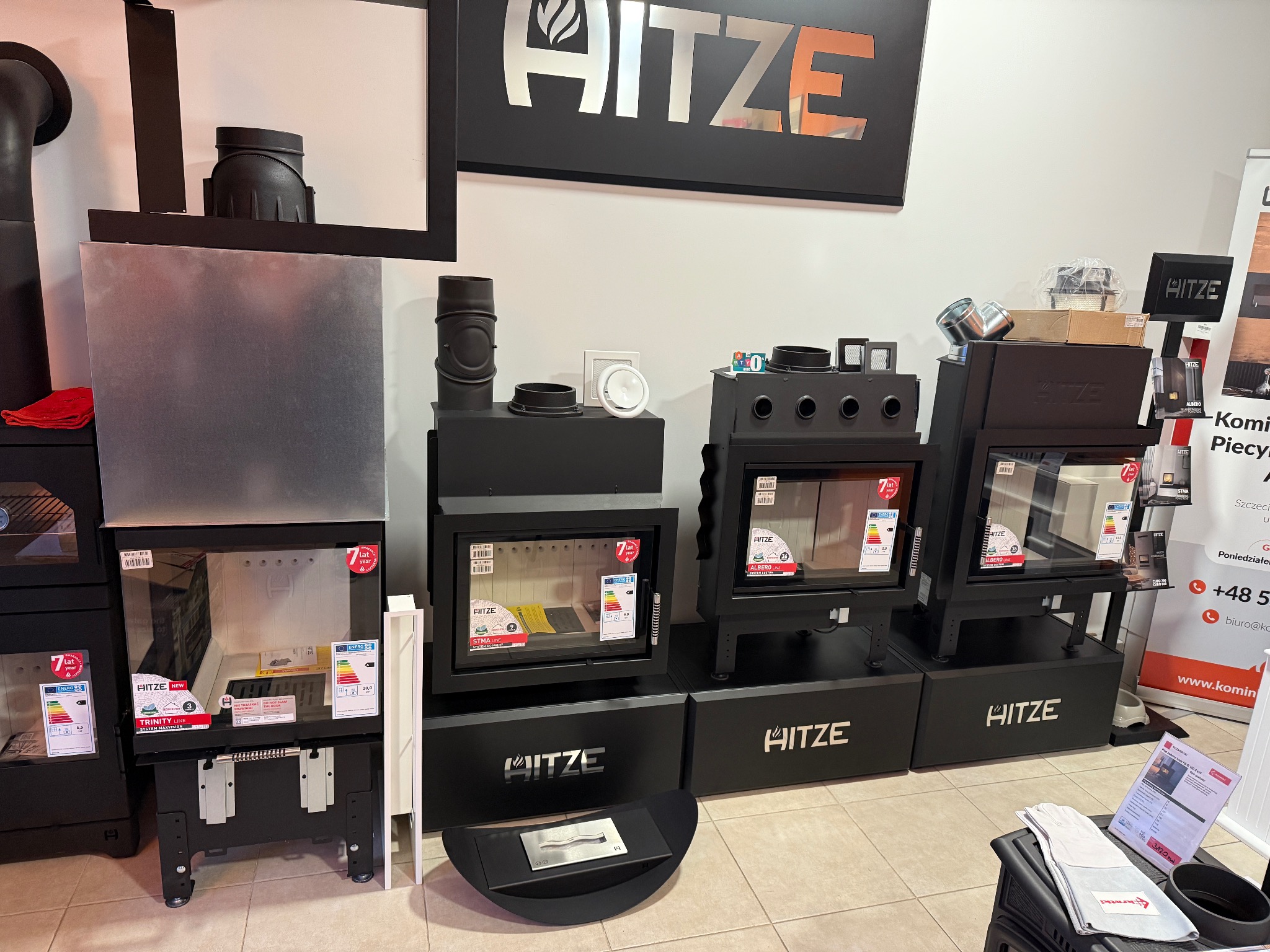 Ekspozycja modeli wkładów kominkowych marki Hitze w salonie, widoczne różne modele z szybami, obudowami i elementami kominowymi, z etykietami energetycznymi i informacjami o produkcie.