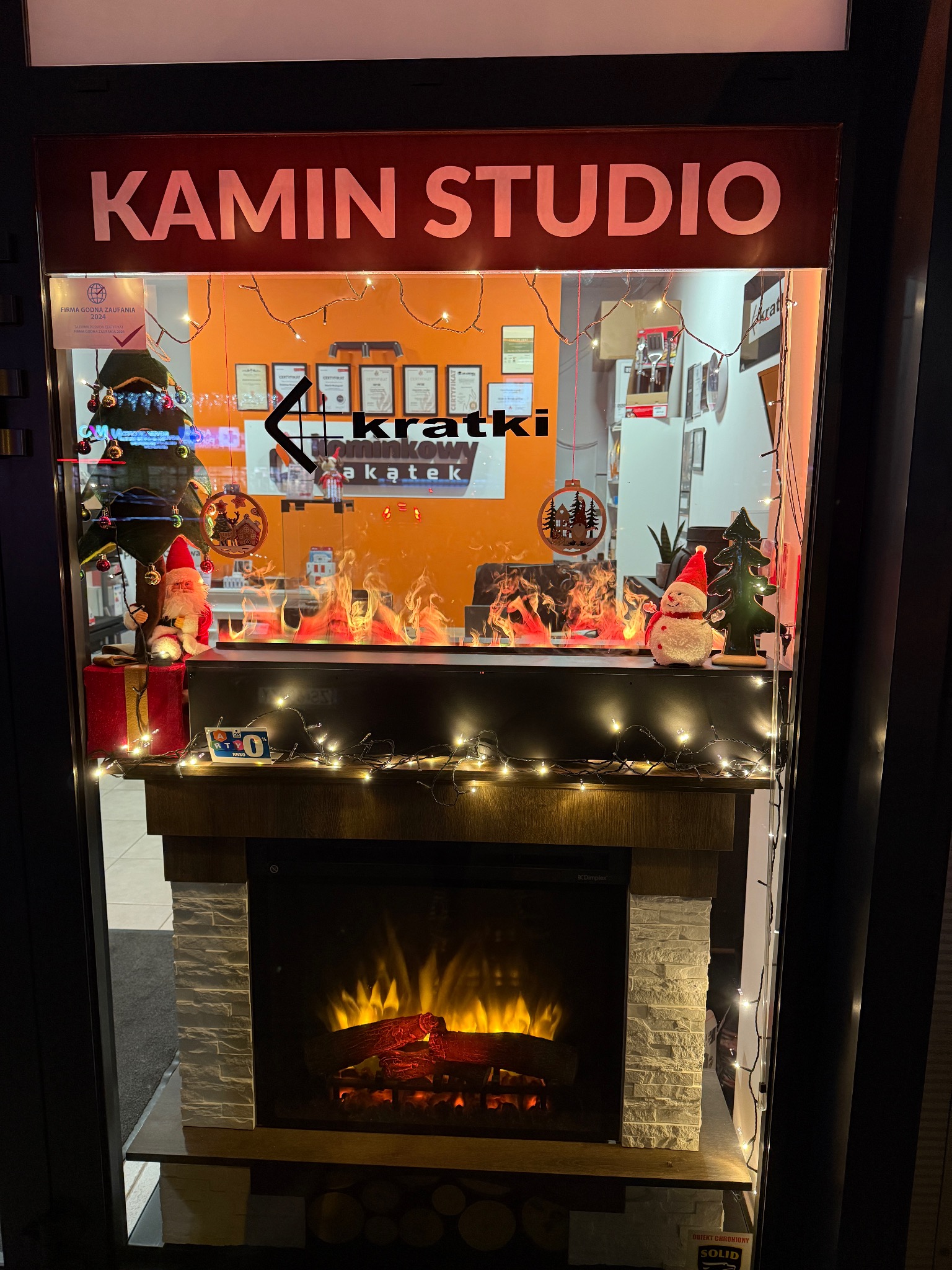 Wystawa salonu kominkowego z elektrycznym kominkiem udekorowanym świątecznymi ozdobami, w tle widoczne certyfikaty i logo firmy.