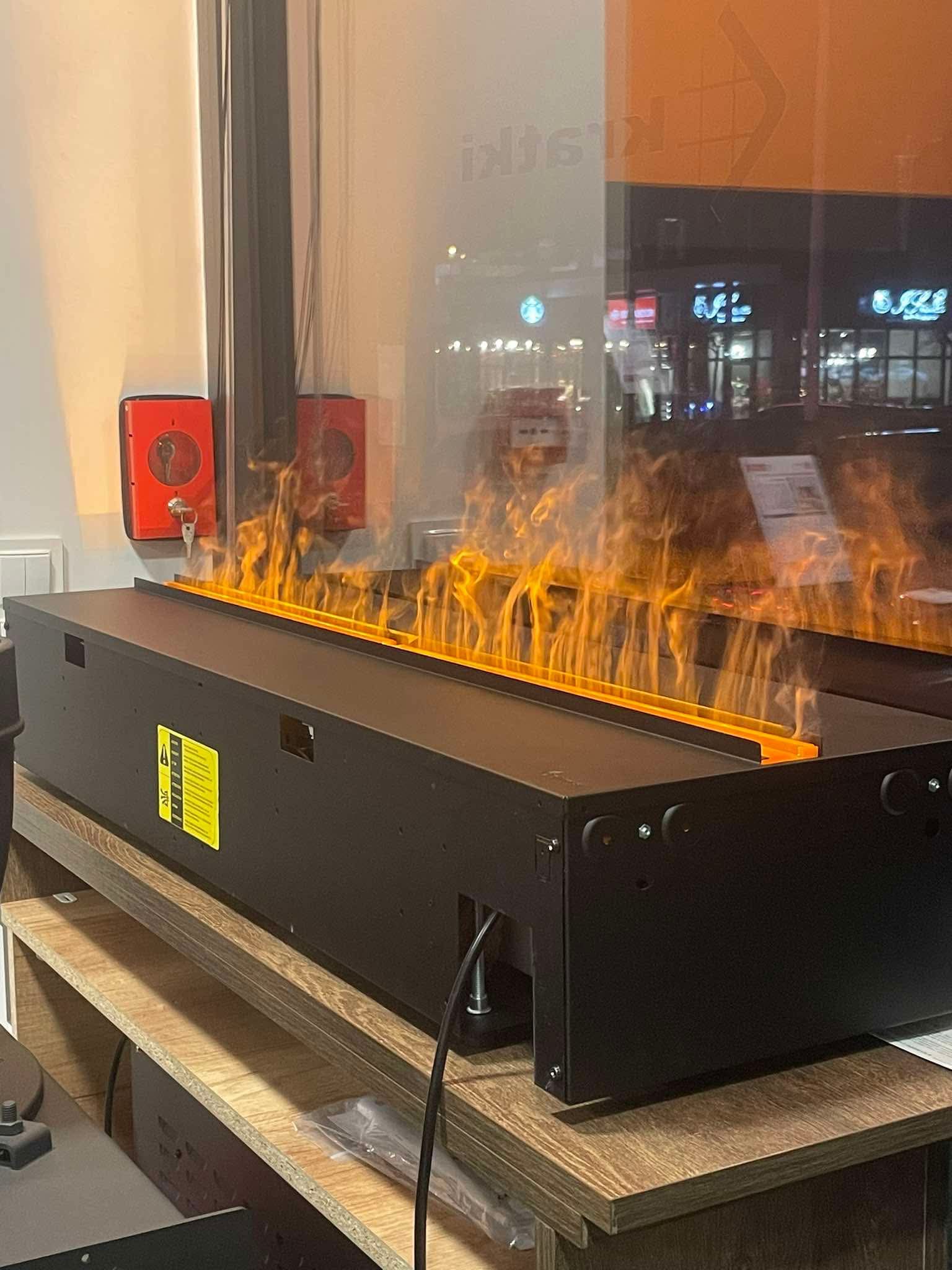 Biokominek w czarnej obudowie, z widocznym płomieniem, umieszczony na drewnianej półce, z odbiciem zewnętrznego otoczenia w szybie.