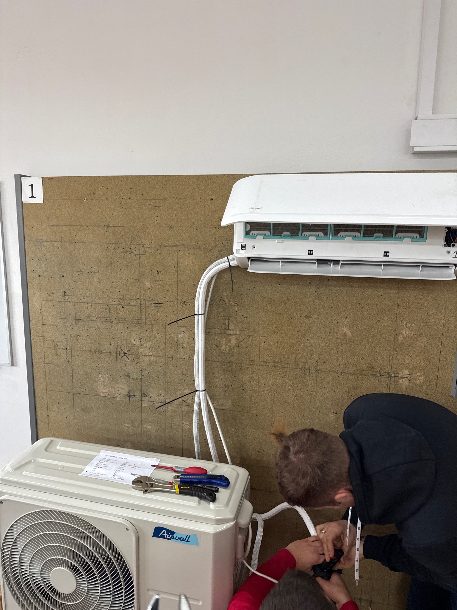Instalacja klimatyzacji Airwell: monterzy podłączają przewody, na agregacie leżą narzędzia i specyfikacja techniczna.