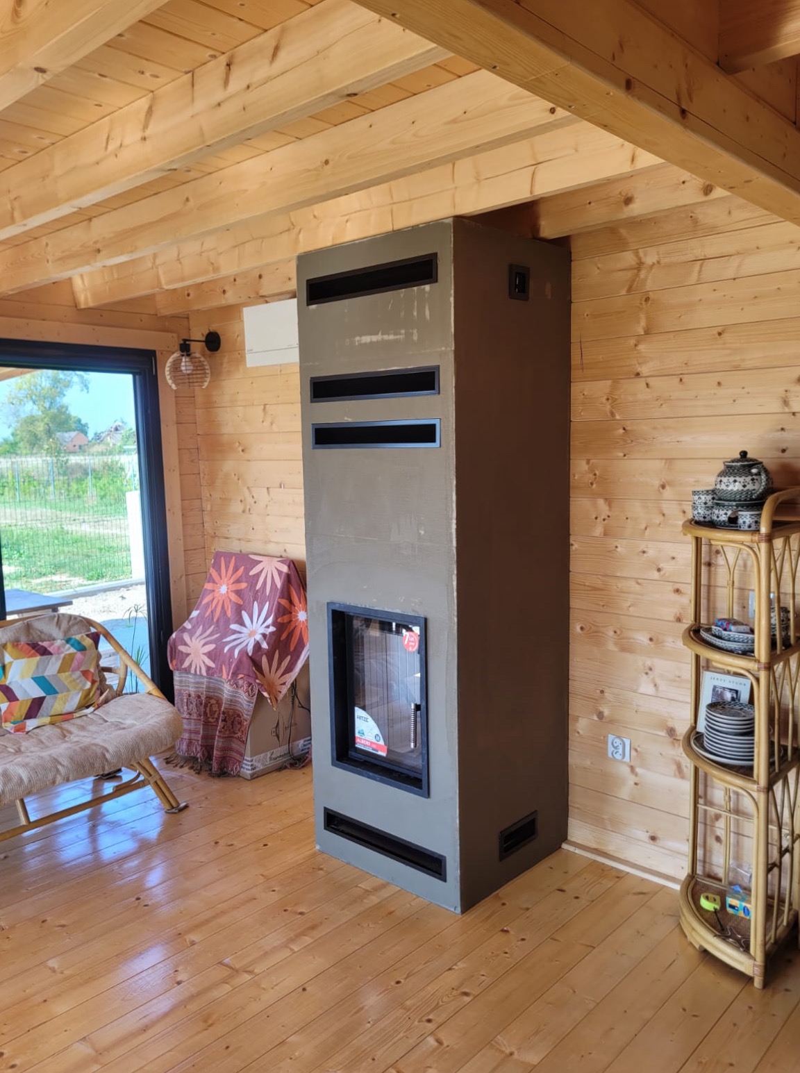 Nowoczesny kominek w obudowie z szarego betonu z czarnymi wstawkami, umieszczony w drewnianym domku, widoczne kratki wentylacyjne, obok regał z dekoracjami.