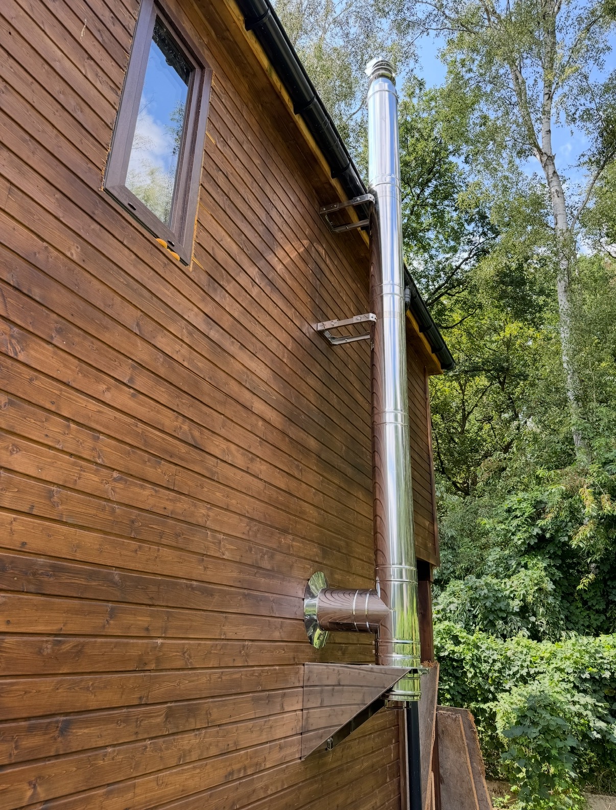 Zewnętrzny, stalowy komin systemowy zamontowany przy drewnianym domu z brązową elewacją, widoczne elementy mocujące.