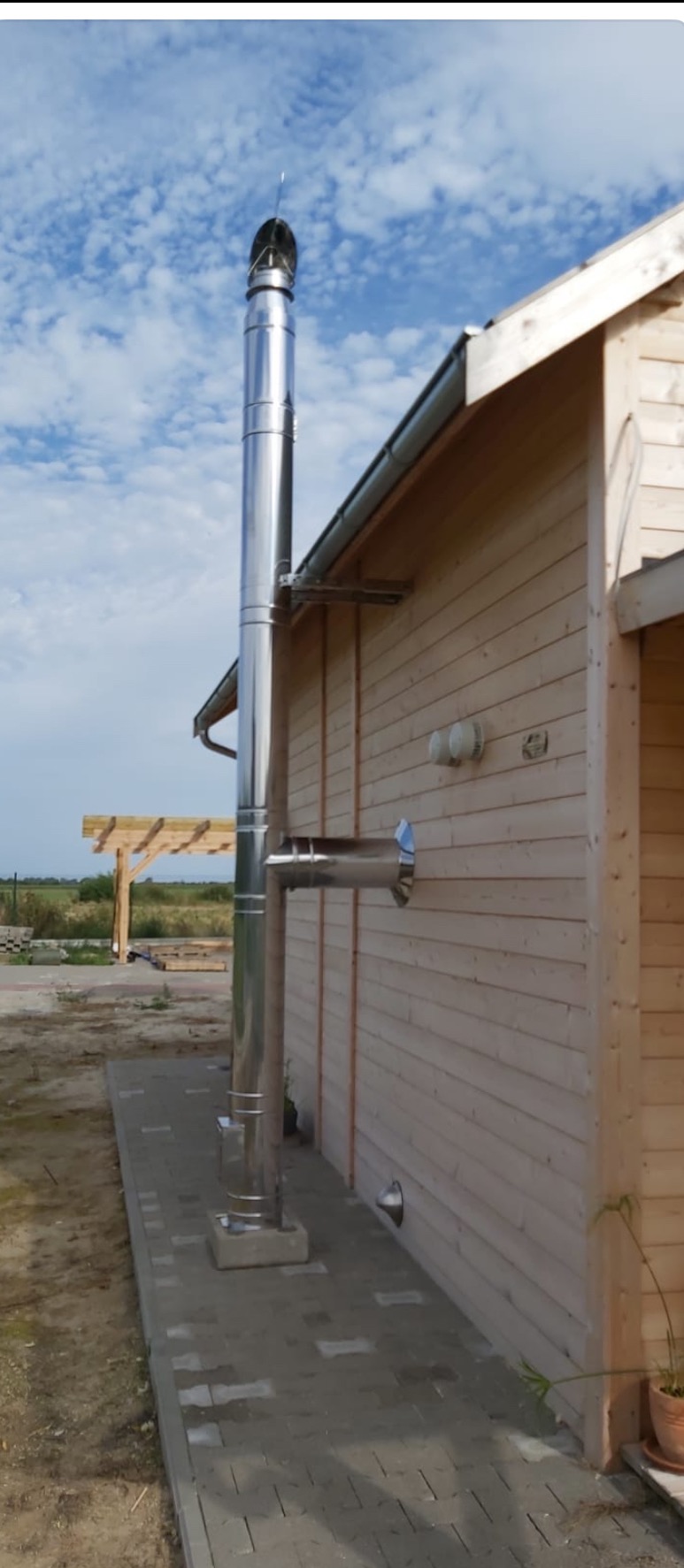 Zewnętrzny, stalowy komin systemowy zamontowany przy drewnianym budynku z jasną elewacją, widoczny fragment chodnika z kostki brukowej i fragment pergoli w tle.