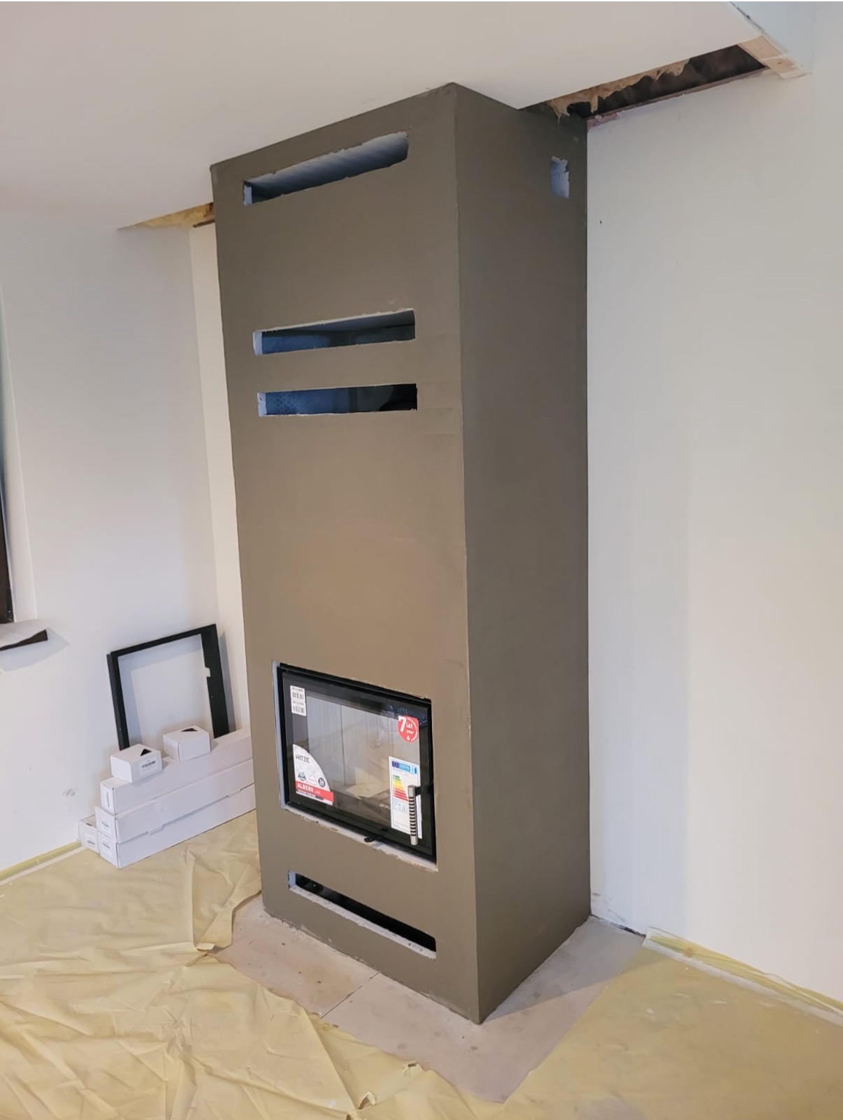 Surowy kominek z szarego betonu z prostokątnym wkładem kominkowym, widoczne otwory wentylacyjne, zabezpieczona folią podłoga i materiały budowlane obok, na etapie wykończenia.