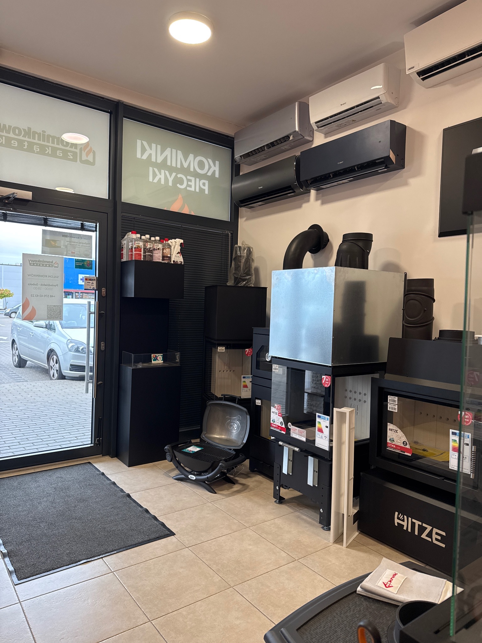 Wnętrze salonu z ekspozycją pieców i kominków, widoczne modele HITZ, rury kominowe, klimatyzatory na ścianie i witryna sklepu z napisem 'Kominki Piece'.