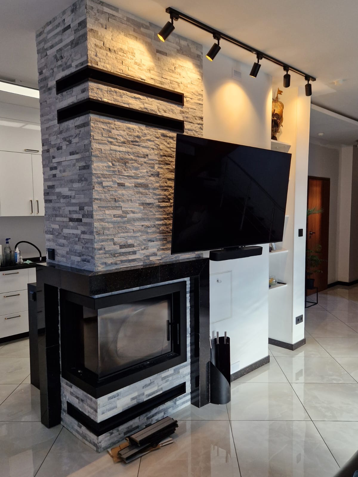 Nowoczesny kominek narożny obudowany szarą cegłą dekoracyjną, z czarnym obramowaniem i blatem z ciemnego granitu, wkomponowany w ścianę z telewizorem i oświetleniem punktowym