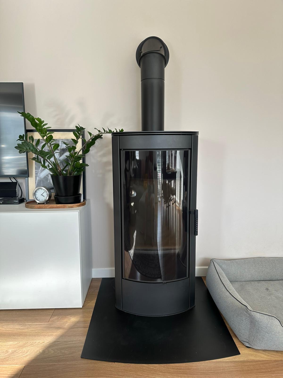 Czarny, nowoczesny kominek wolnostojący z szybą, umieszczony na czarnej macie ochronnej, obok legowiska dla psa i szafki z rośliną doniczkową.
