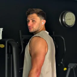 Mężczyzna w sportowej koszulce bez rękawów, z słuchawkami bezprzewodowymi w uchu, odwraca się w siłowni, w tle czarna ściana i zegar.