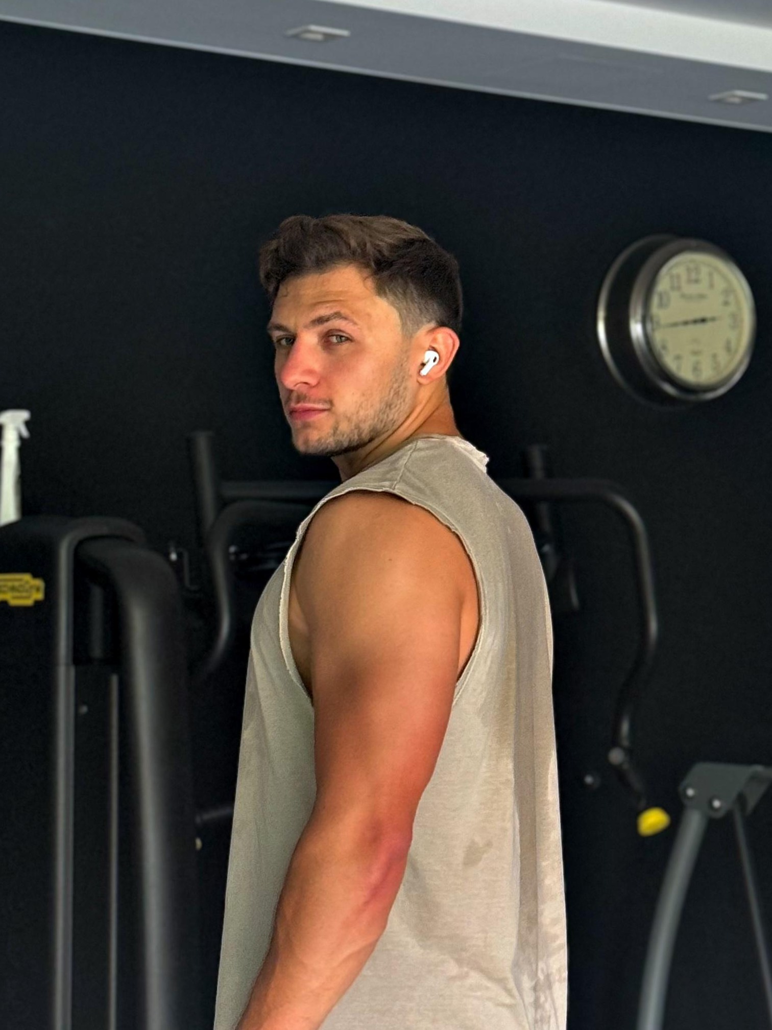 Mężczyzna w sportowej koszulce bez rękawów, z słuchawkami bezprzewodowymi w uchu, odwraca się w siłowni, w tle czarna ściana i zegar.