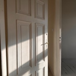 Patrycja Kaczmarek Podwójna Agentka - Otwarta, biała, drewniana, wewnętrzna, kasetonowa, częściowo oświetlona drzwiami w pustym pokoju z bieloną podłogą.