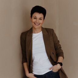 Patrycja Kaczmarek Podwójna Agentka - Uśmiechnięta kobieta w brązowej marynarce i jeansach, oparta o beżową ścianę, z zegarkiem na nadgarstku.