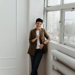 Patrycja Kaczmarek Podwójna Agentka - Uśmiechnięta kobieta w brązowej marynarce, oparta o białą ścianę z grzejnikiem i oknem z widokiem na budynki w tle, trzymająca okulary w dłoni.