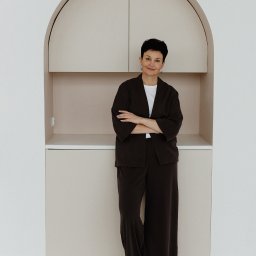 Patrycja Kaczmarek Podwójna Agentka - Kobieta z krótkimi włosami, ubrana w ciemny garnitur, stoi na tle zabudowy meblowej w kształcie łuku, z beżowymi szafkami i półkami.