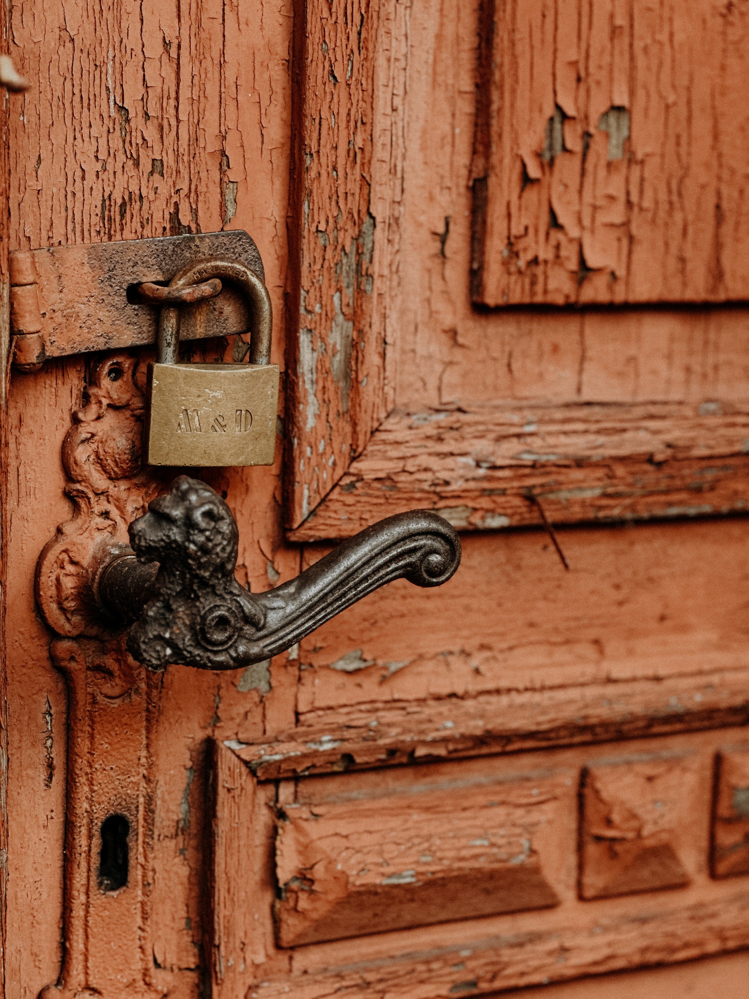 Zardzewiały kłódka z napisem 'M&D' zapięta na starej, pomarańczowej, drewnianej furtce z łuszczącą się farbą i ozdobną, żeliwną klamką w kształcie głowy zwierzęcia.