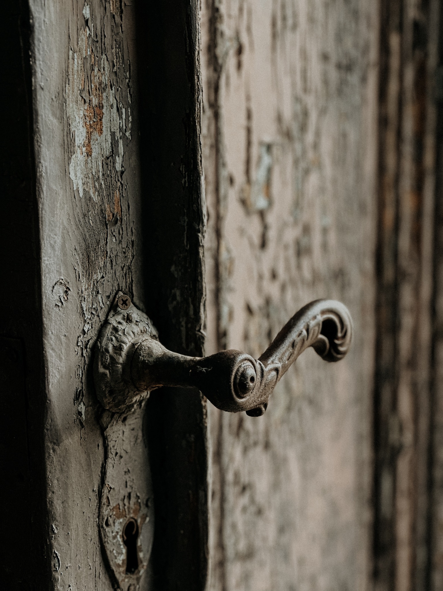 Zardzewiała, ozdobna klamka i zamek w starych, drewnianych drzwiach z łuszczącą się farbą, widoczne pionowe deski.