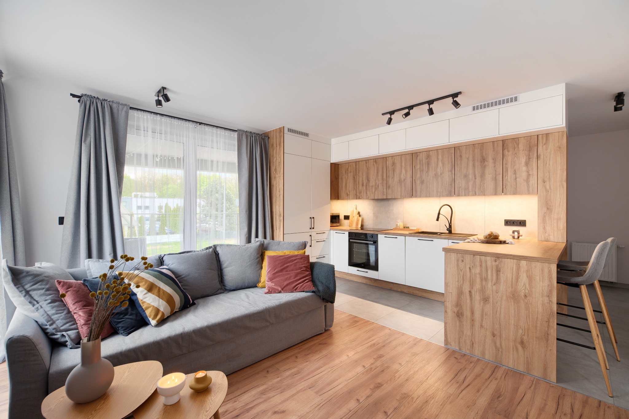 Jasne, nowoczesne wnętrze mieszkania z szarą sofą, drewnianą podłogą i kuchnią z drewnianymi akcentami, oświetloną lampami sufitowymi.