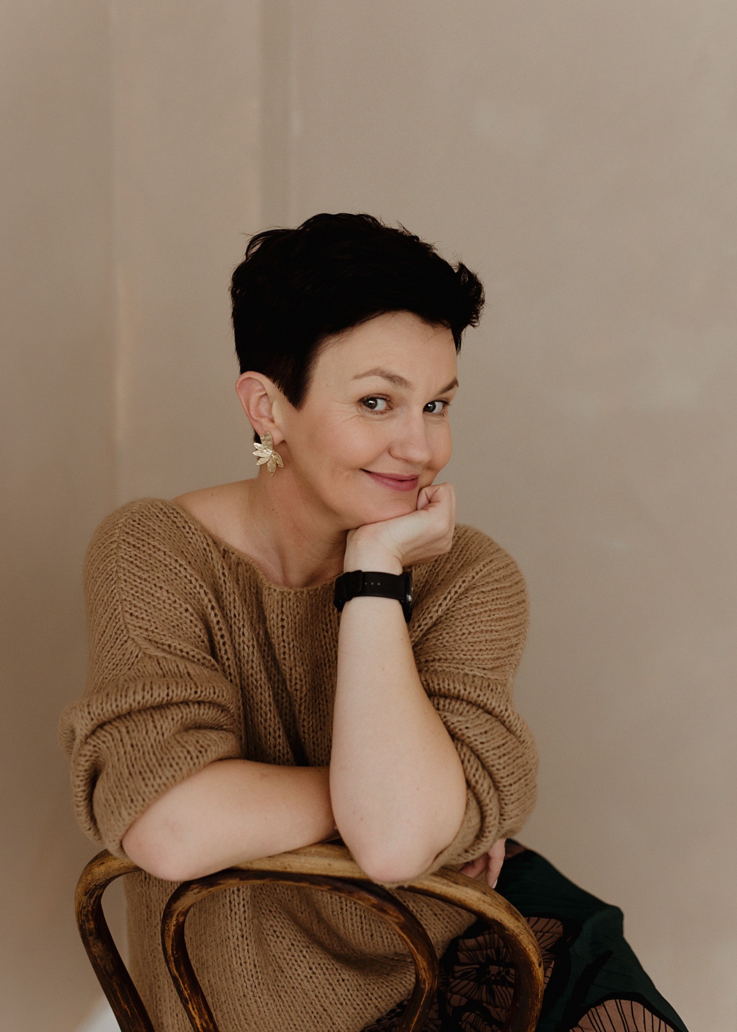 Portret uśmiechniętej kobiety z krótkimi, ciemnymi włosami, w beżowym swetrze, opierającej podbródek na dłoni z czarnym zegarkiem, siedzącej na drewnianym krześle z dekoracyjnymi kolczykami...
