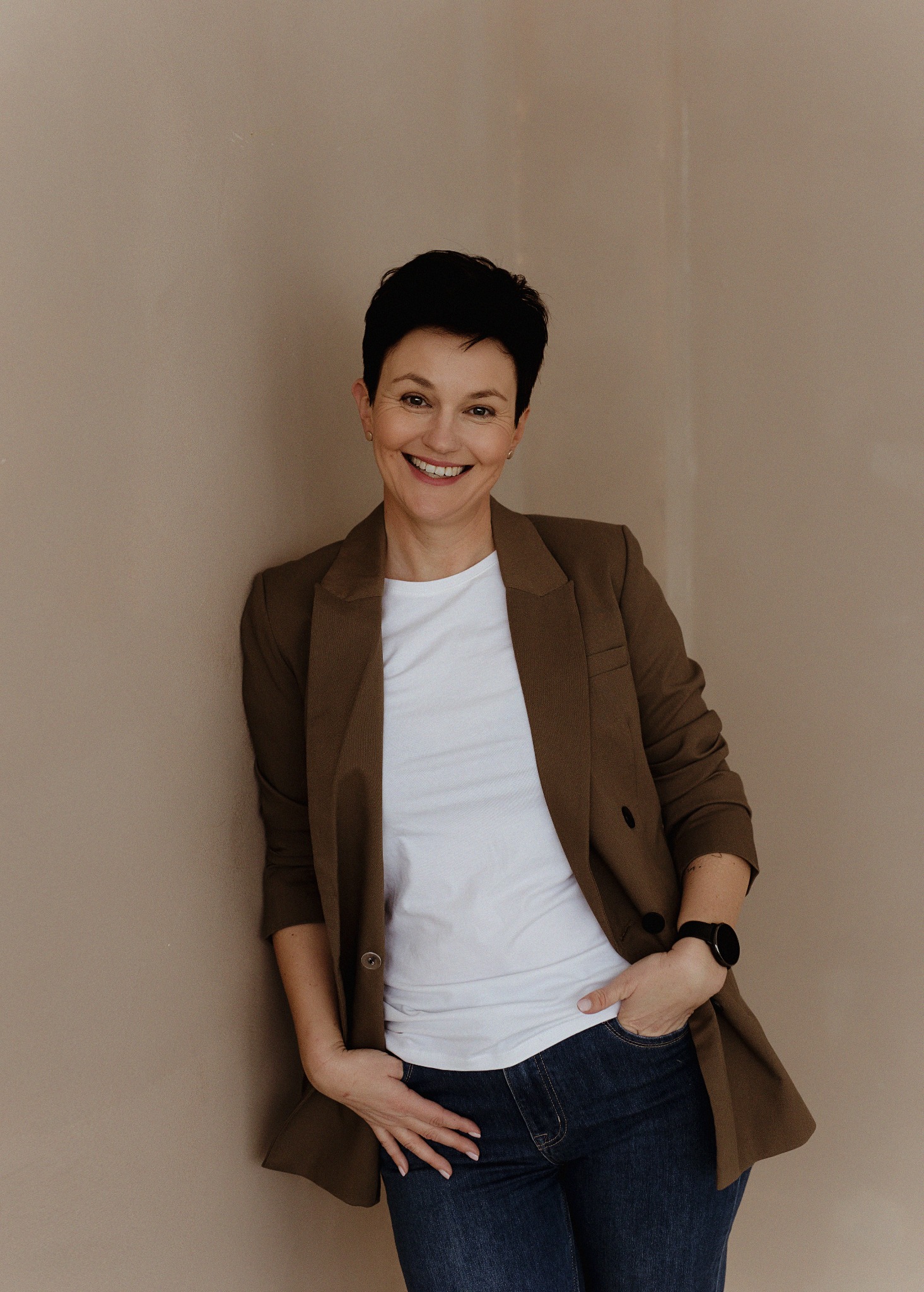 Uśmiechnięta kobieta w brązowej marynarce i jeansach, oparta o beżową ścianę, z zegarkiem na nadgarstku.