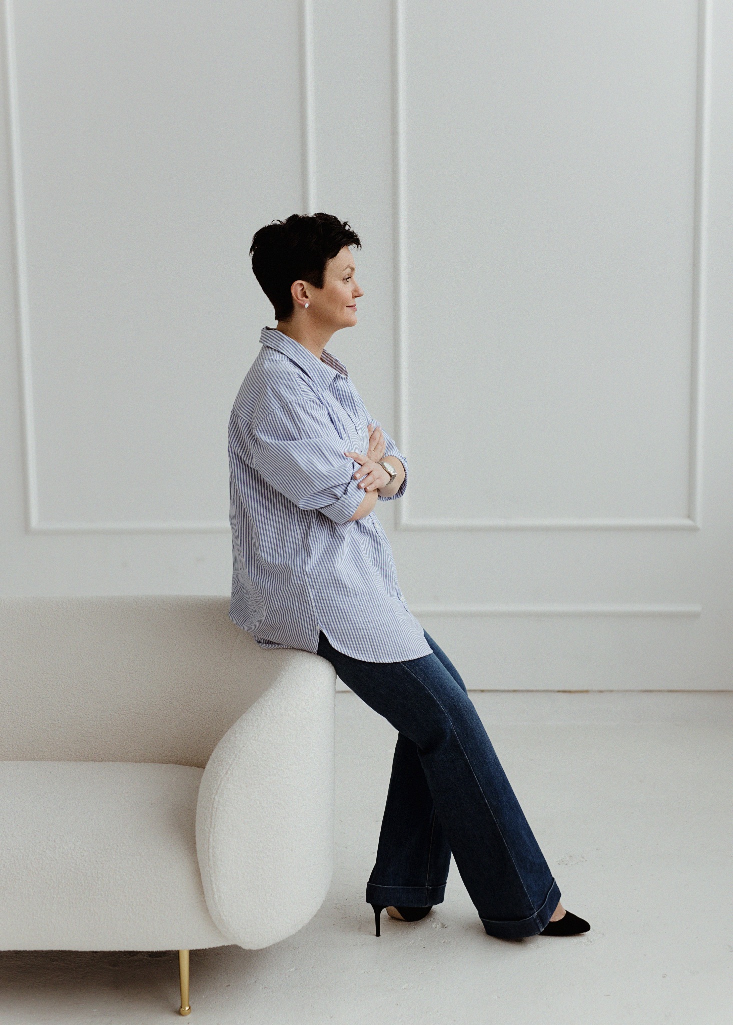 Portret kobiety w niebieskiej koszuli w paski, siedzącej na białej sofie z nogami w ciemnych jeansach i czarnych butach na obcasie, na tle białej ściany z dekoracyjnymi panelami.