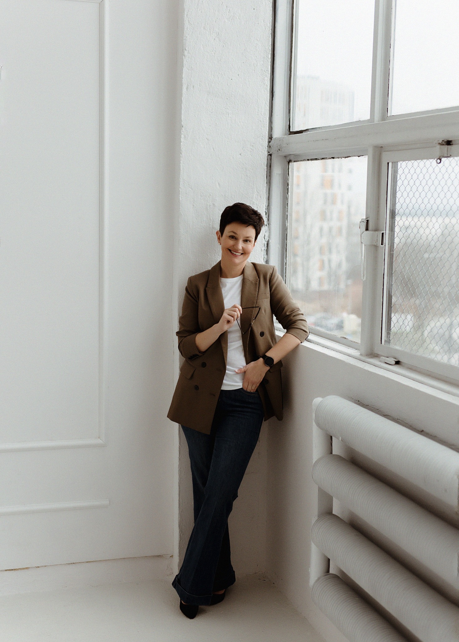 Uśmiechnięta kobieta w brązowej marynarce, oparta o białą ścianę z grzejnikiem i oknem z widokiem na budynki w tle, trzymająca okulary w dłoni.