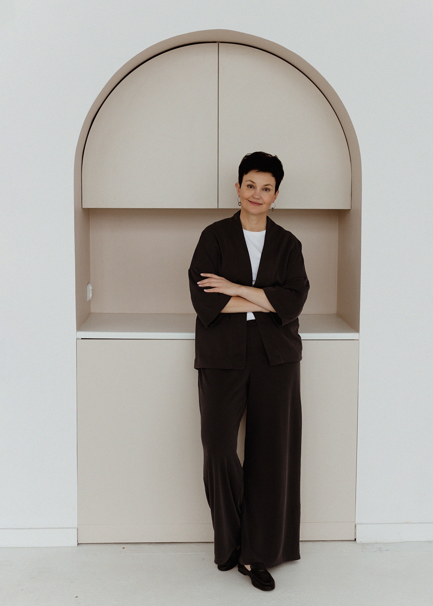 Kobieta z krótkimi włosami, ubrana w ciemny garnitur, stoi na tle zabudowy meblowej w kształcie łuku, z beżowymi szafkami i półkami.