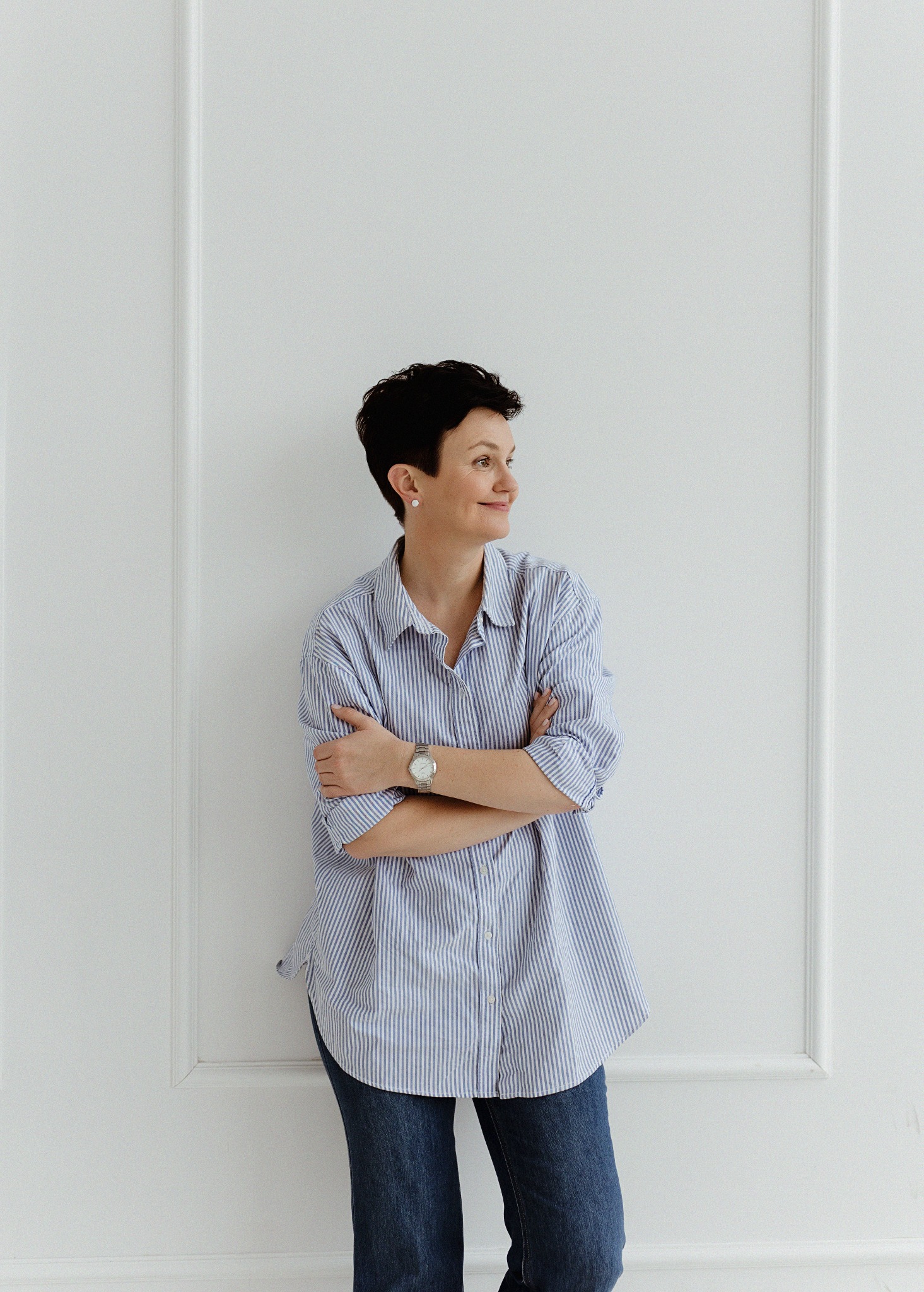 Uśmiechnięta kobieta z krótkimi, ciemnymi włosami, ubrana w niebieską koszulę w paski i dżinsy, stoi na tle białej ściany z dekoracyjnymi panelami, spoglądając w bok.