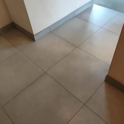 A&S BUD ADRIAN SZCZYGIEŁ - Jasne, kwadratowe płytki podłogowe z szarą listwą przypodłogową i fragmentem buta w rogu. W tle widoczne białe ściany i fragment pokoju.