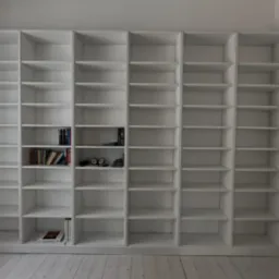 Biała, zabudowana biblioteczka z półkami, częściowo wypełniona książkami i dekoracjami, na tle jasnej ściany i podłogi z desek.