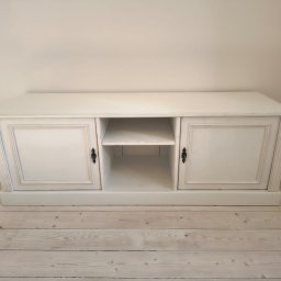 A&S BUD ADRIAN SZCZYGIEŁ - Biała, postarzana szafka RTV z dwoma zamykanymi szafkami i otwartą półką pośrodku, na jasnej drewnianej podłodze, w jasnym wnętrzu.