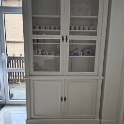 A&S BUD ADRIAN SZCZYGIEŁ - Biały, dwuczęściowy kredens z przeszkloną górą i pełnymi drzwiczkami na dole, wypełniony naczyniami i akcesoriami, ustawiony w jasnym wnętrzu obok okna.