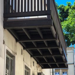 A&S BUD ADRIAN SZCZYGIEŁ - Ciemnobrązowy drewniany balkon z widoczną kratownicową konstrukcją od spodu, zamontowany na jasnej, lekko zniszczonej elewacji starego budynku z klimatyzatorem.