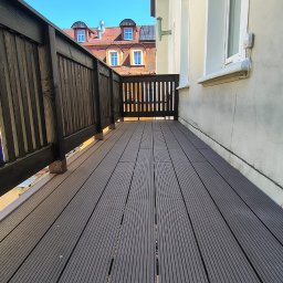 A&S BUD ADRIAN SZCZYGIEŁ - Nowo ułożony ciemnoszary taras kompozytowy na balkonie, z ciemną drewnianą balustradą, wzdłuż białej ściany budynku. W tle widoczne czerwone dachy z lukarnami pod błękitnym niebem.