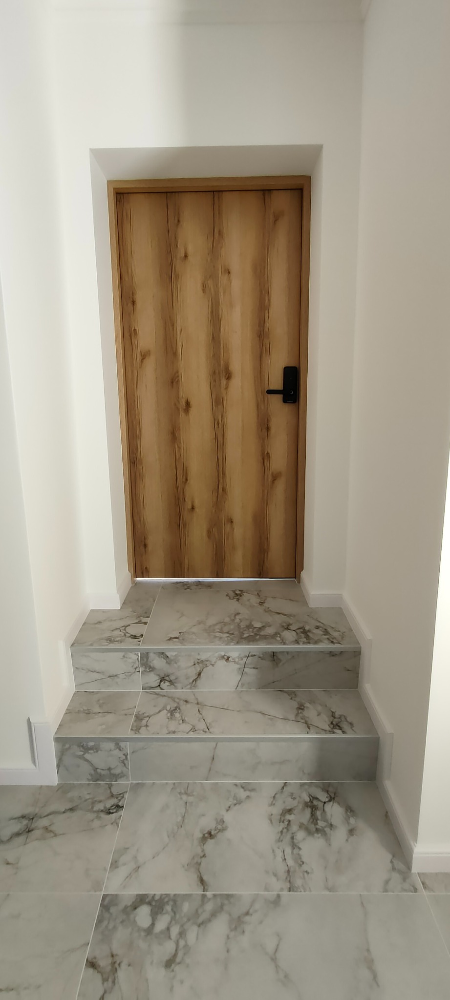 Nowoczesne wykończenie wnętrza: drewniane drzwi z czarną klamką i marmurowe schody w jasnym pomieszczeniu. Minimalizm i elegancja w detalu.