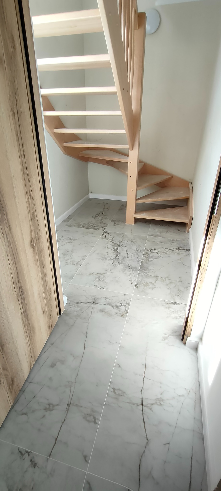 Jasne drewniane schody w małym przedpokoju z szarymi płytkami imitującymi marmur. Widoczne drzwi po bokach, białe ściany i sufit z lampą.