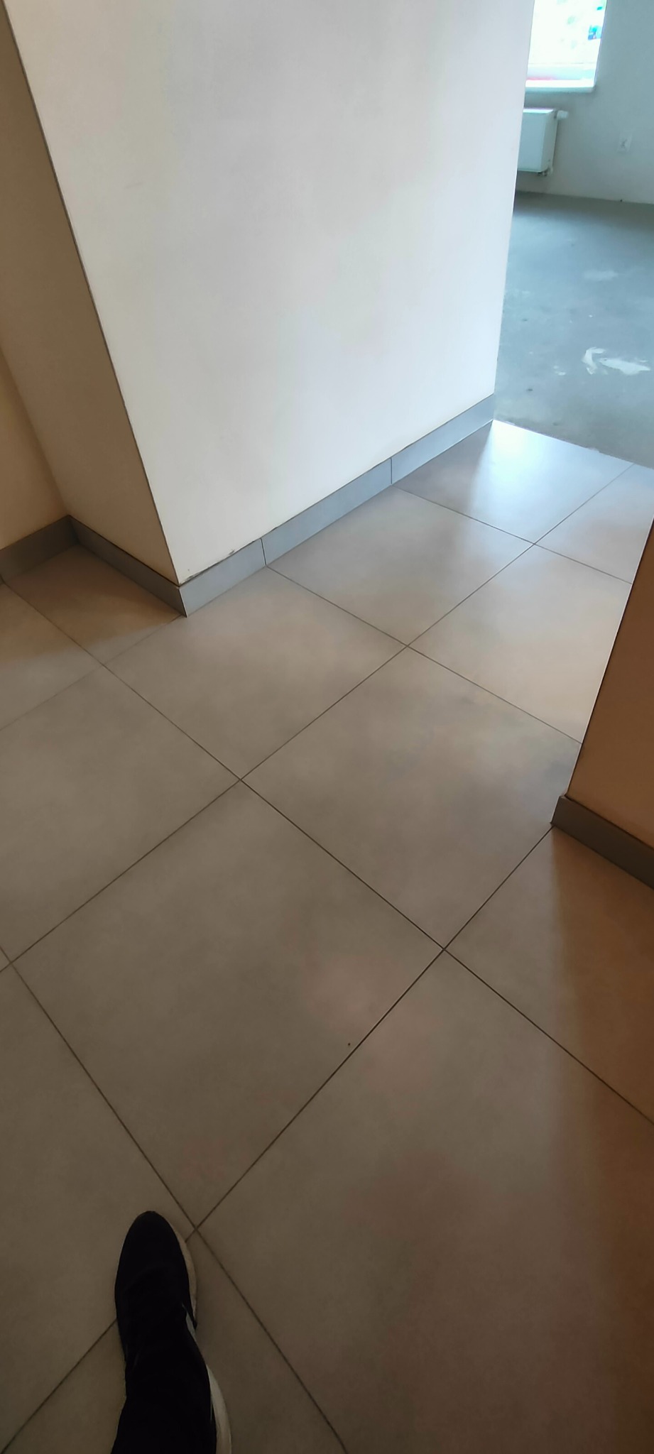 Jasne, kwadratowe płytki podłogowe z szarą listwą przypodłogową i fragmentem buta w rogu. W tle widoczne białe ściany i fragment pokoju.