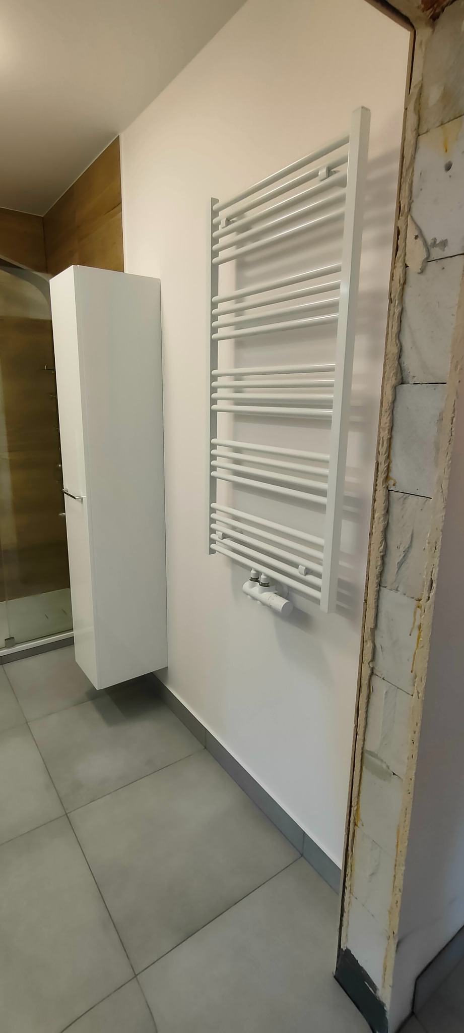 Biała szafka łazienkowa, grzejnik drabinkowy na ścianie i szara podłoga z płytek w łazience. Fragment ściany w trakcie remontu.