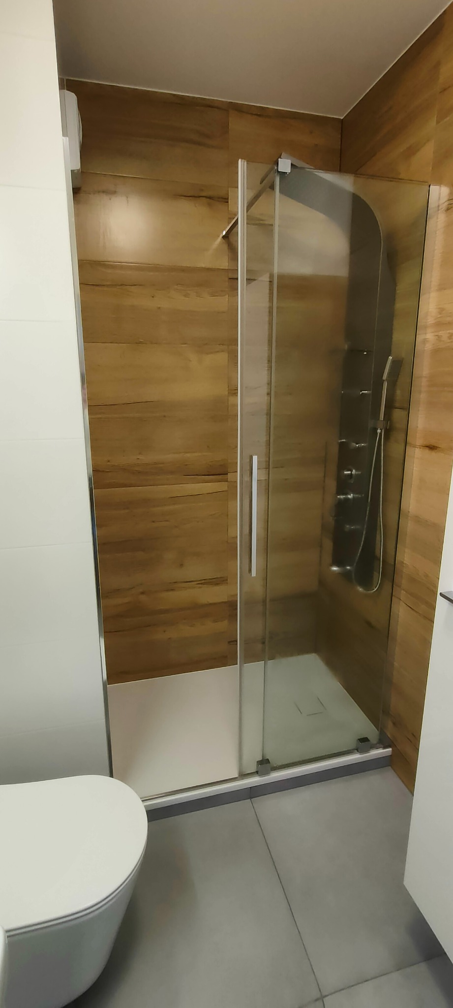 Nowoczesna kabina prysznicowa z drewnopodobnymi płytkami na ścianie. Szklana ścianka z metalowymi okuciami. Widoczna toaleta i szara podłoga.