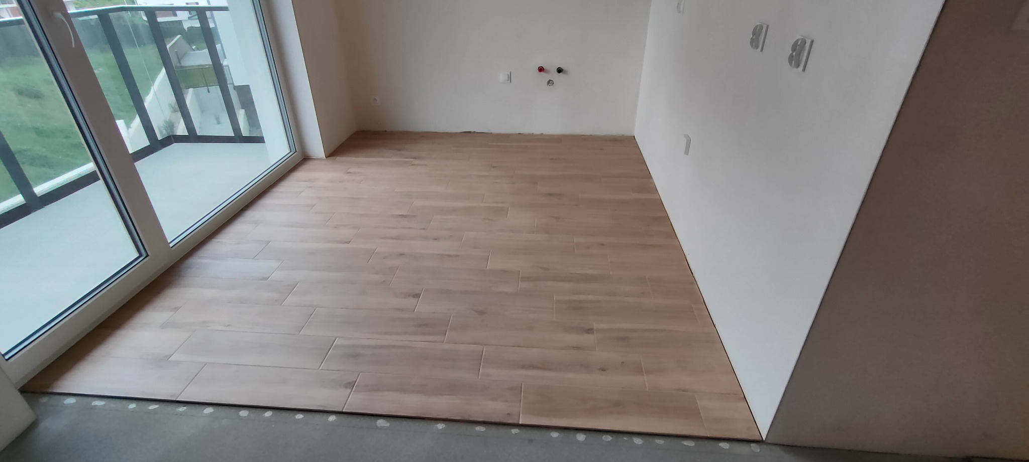Ułożona podłoga z płytek imitujących drewno w jasnym wnętrzu z dużym oknem balkonowym. Widoczne fragmenty instalacji elektrycznej na ścianie.