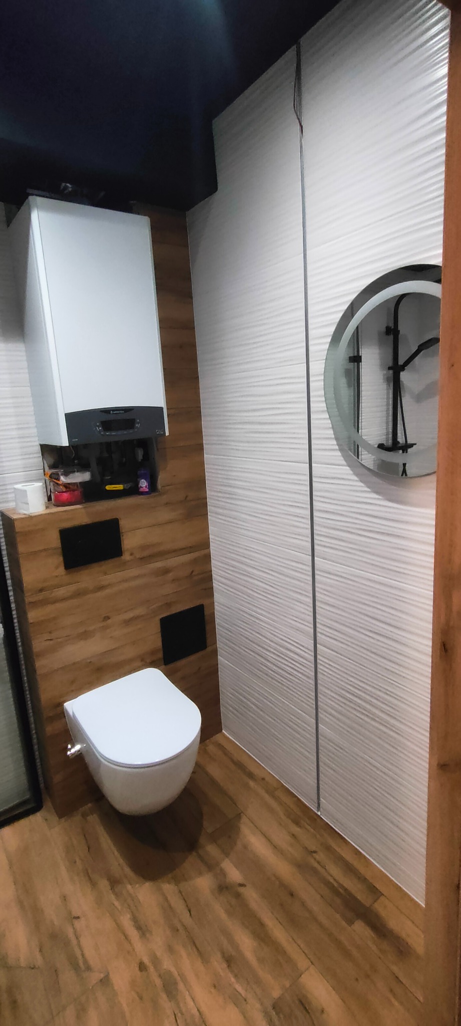 Nowoczesna łazienka z podwieszaną toaletą, drewnopodobnymi płytkami na podłodze i ścianie, oraz okrągłym lustrem z podświetleniem LED.