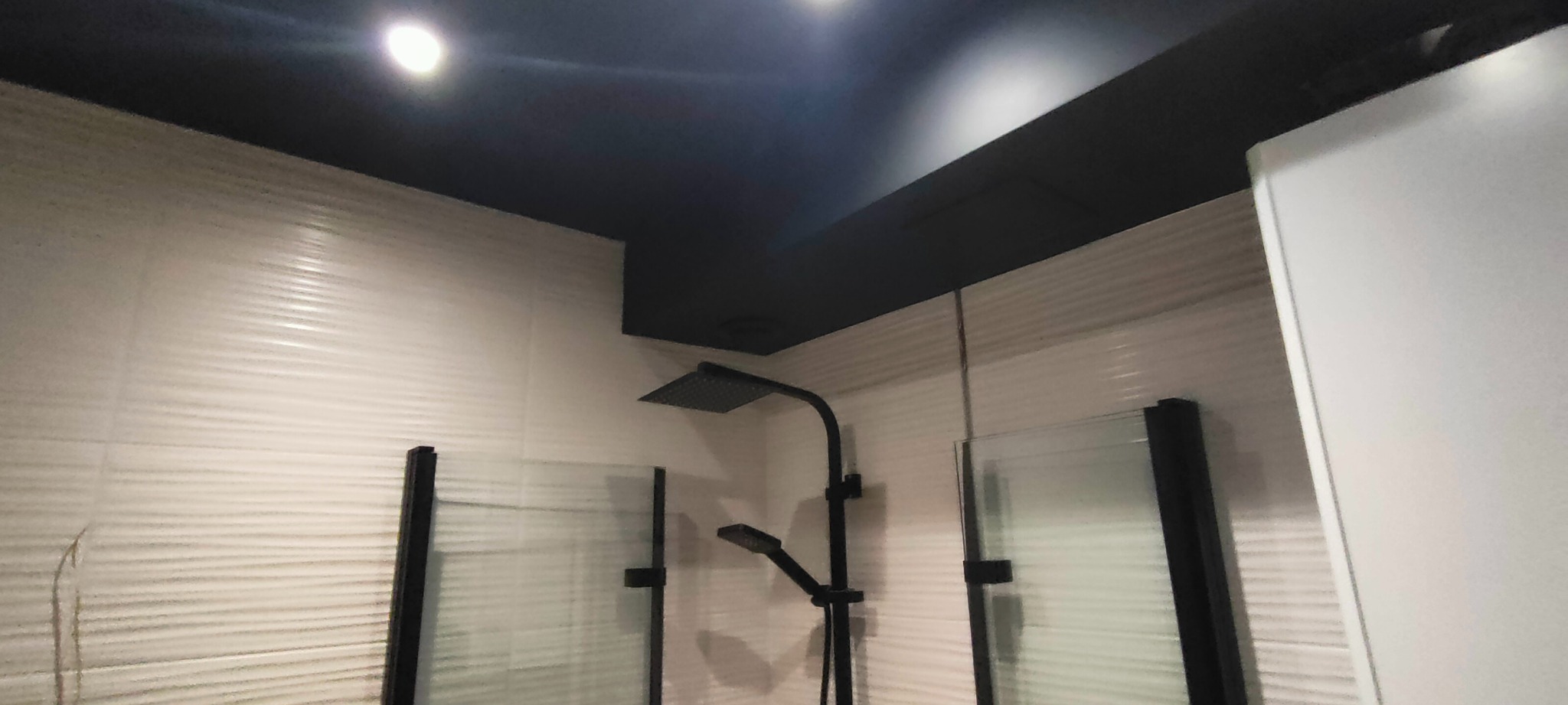 Nowoczesna łazienka z czarnym sufitem, armaturą i białymi płytkami z poziomym wzorem. Kabina prysznicowa z matowego szkła. Oświetlenie punktowe.