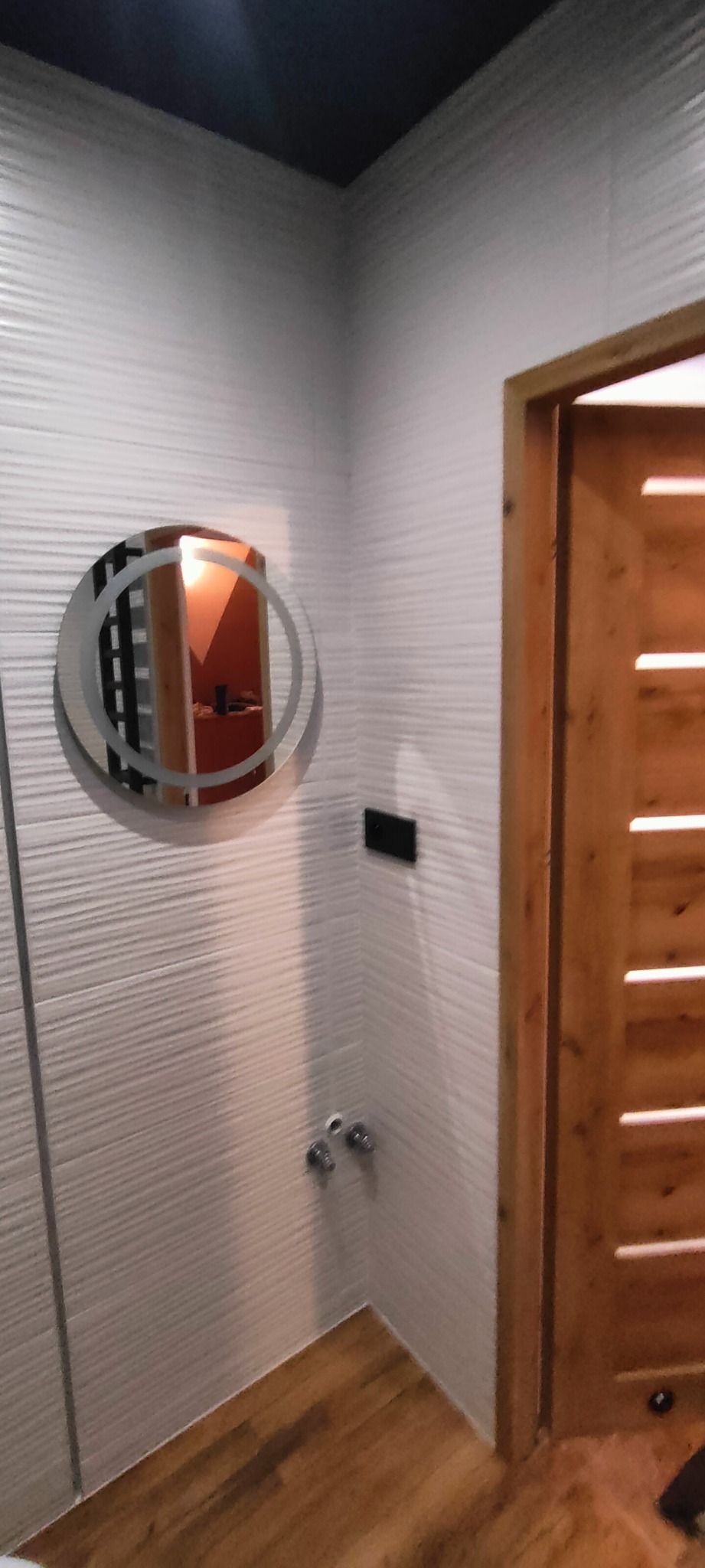 Wykończona łazienka z okrągłym lustrem LED, teksturowanymi białymi płytkami ściennymi i drewnianymi drzwiami z poziomymi szczelinami. Nowoczesny design.