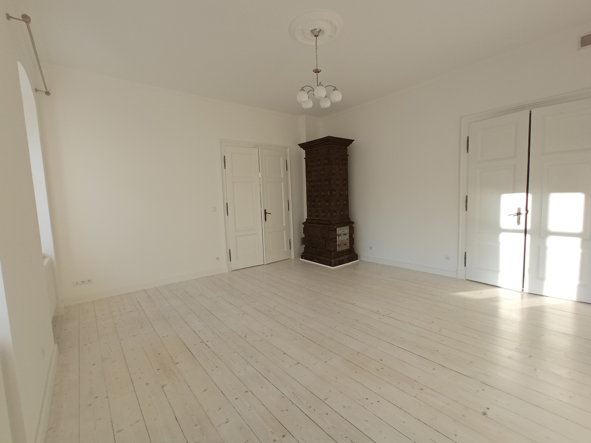 Jasne, przestronne wnętrze z bielonymi deskami podłogowymi, białymi ścianami i drzwiami. Centralnym punktem jest ciemny, kaflowy piec.