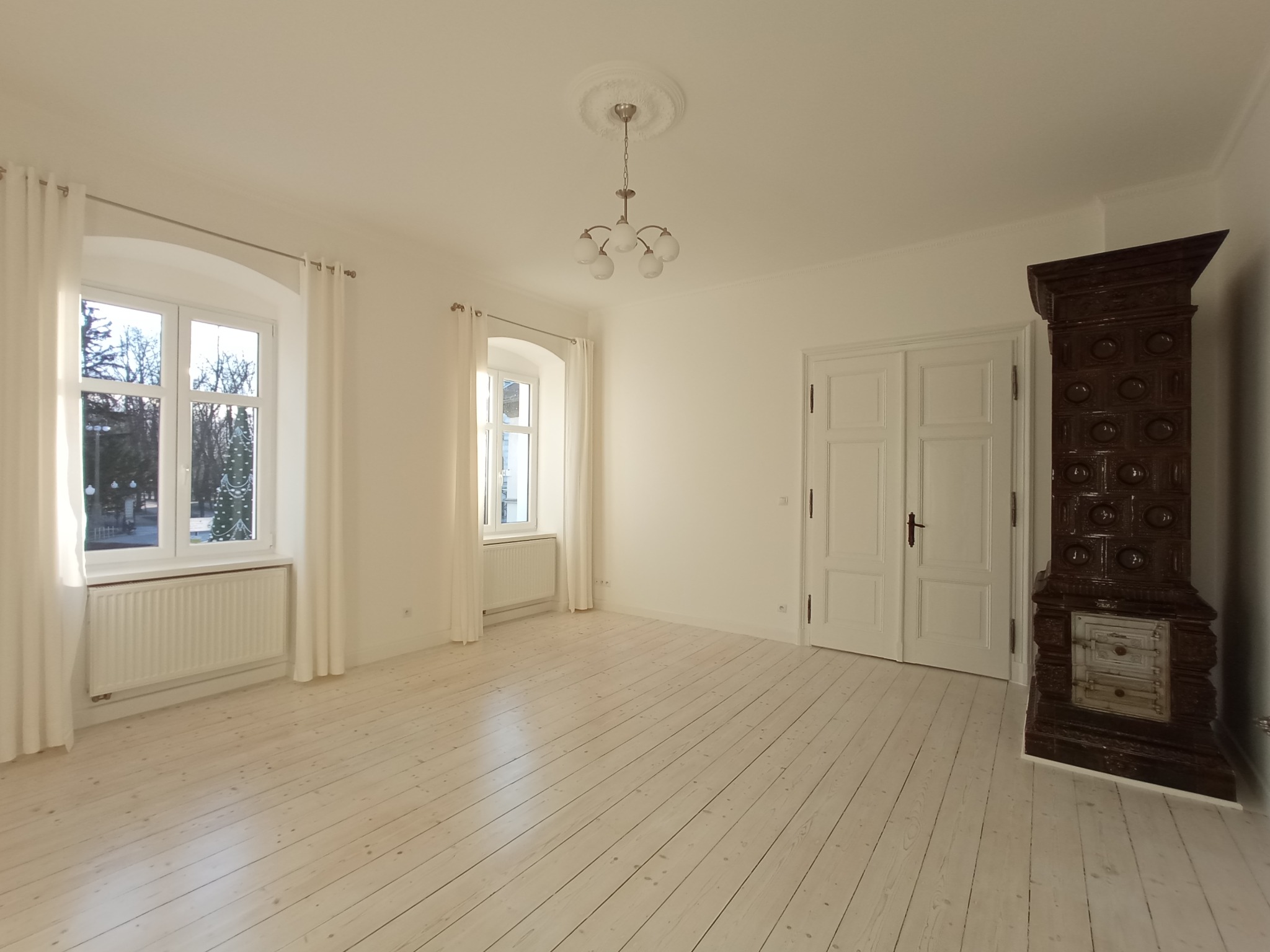 Jasne, przestronne wnętrze z bielonymi deskami podłogowymi, białymi ścianami, oknami z firanami i ozdobnym, ciemnym piecem kaflowym w rogu.