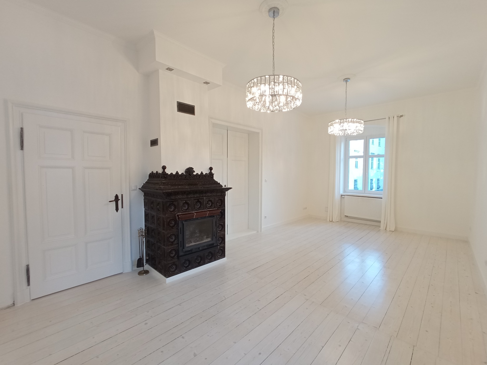 Jasne, przestronne wnętrze z drewnianą podłogą, ozdobnym piecem kaflowym i dwoma kryształowymi żyrandolami. Białe ściany i stolarka okienna.