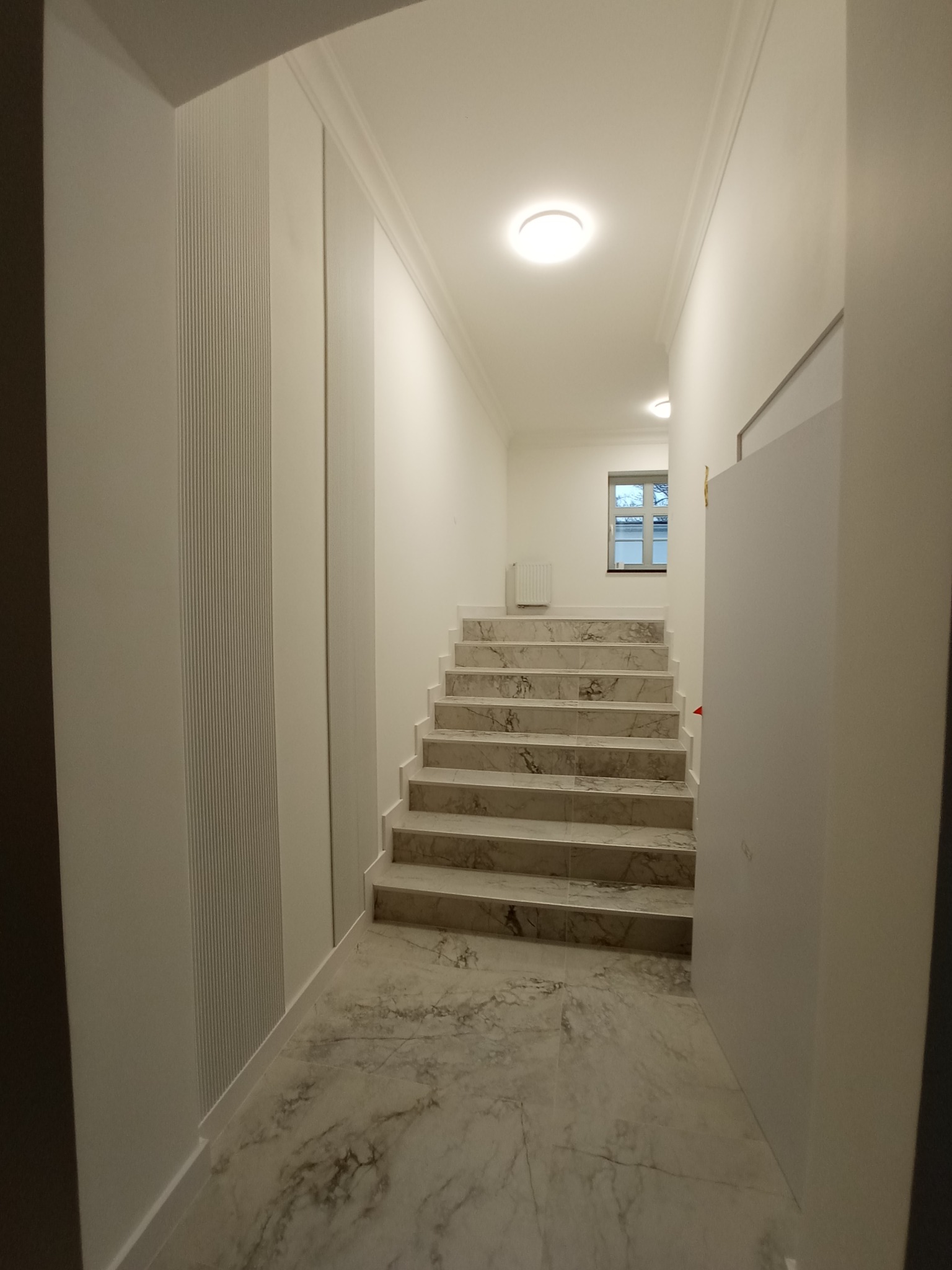 Wykończone marmurowe schody w jasnym korytarzu z dekoracyjną sztukaterią i listwami przypodłogowymi. Widok na okno na końcu korytarza.