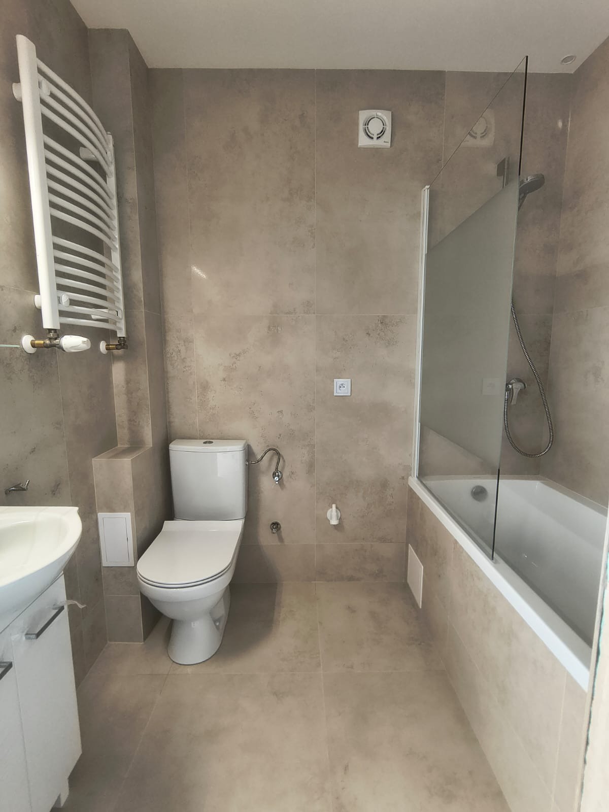 Jasna, beżowa łazienka po remoncie: biała toaleta, wanna z szybą, grzejnik drabinkowy i płytki imitujące kamień. Widok na całe pomieszczenie, neutralne oświetlenie.