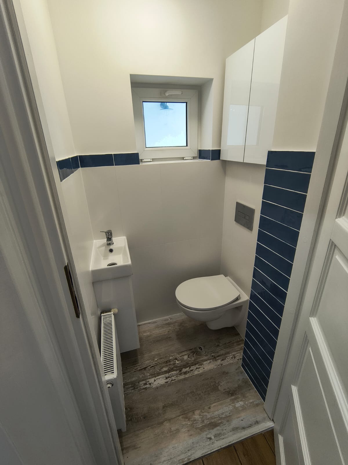 Mała, odnowiona łazienka z białą armaturą, niebieskimi akcentami i drewnopodobną podłogą. Widoczna umywalka, toaleta i grzejnik.