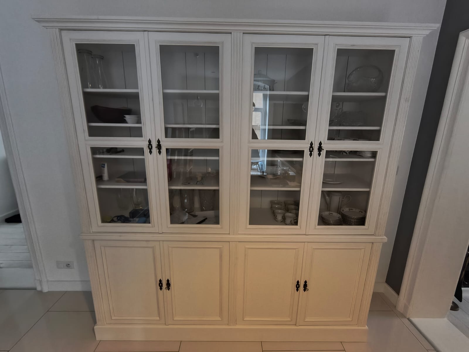 Biały, przeszklony kredens w stylu prowansalskim z ciemnymi uchwytami, wypełniony naczyniami i dekoracjami, ustawiony na jasnej podłodze w pokoju.