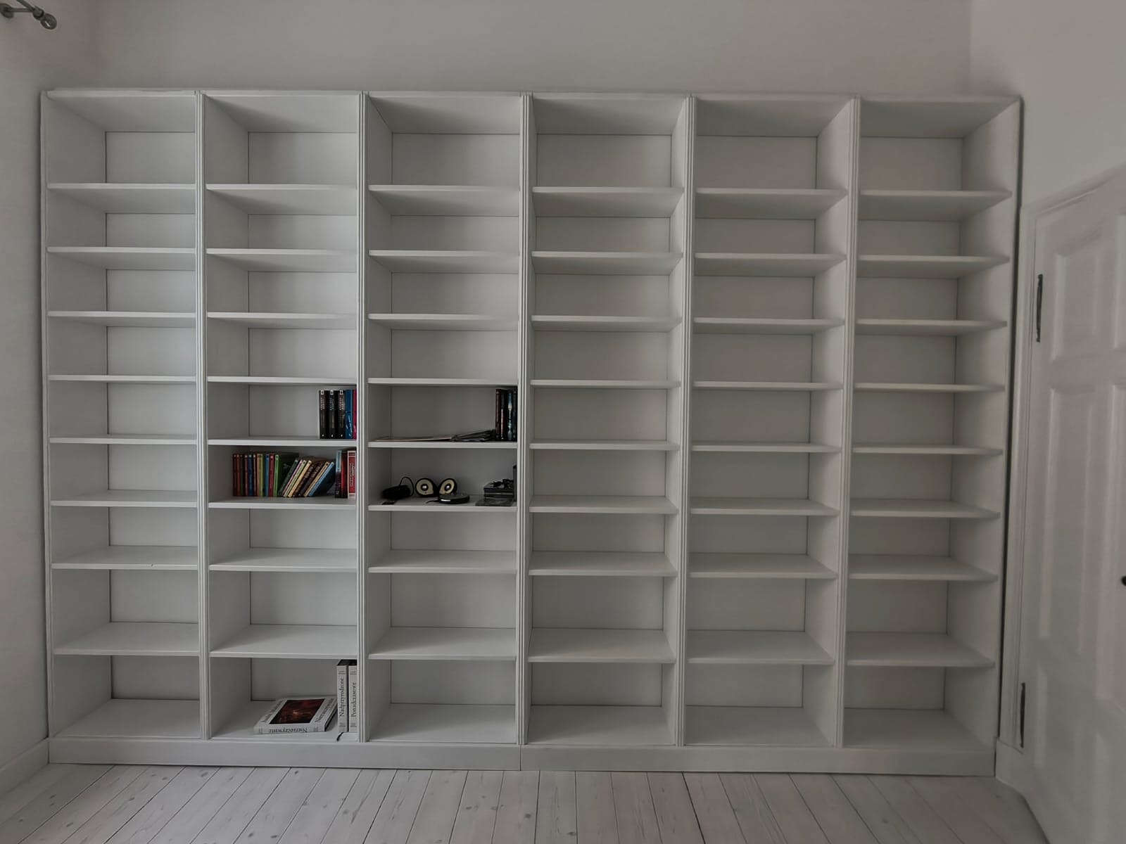 Biała, zabudowana biblioteczka z półkami, częściowo wypełniona książkami i dekoracjami, na tle jasnej ściany i podłogi z desek.